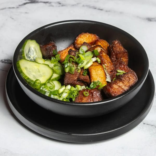 A black bowl of crispy pork belly and potatoes, garnished with sliced cucumbers and green onions.