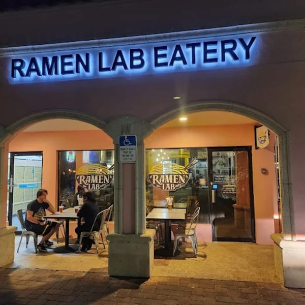 Exterior of Ramen Lab Eatery at night, with a glowing sign and two people dining outside.